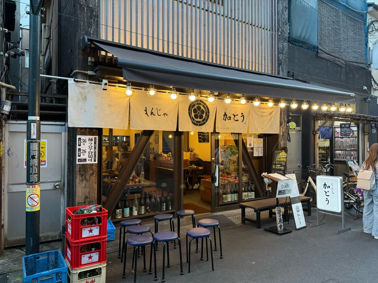 もんじゃ加とうの暖簾と入口、店先に並ぶ丸椅子